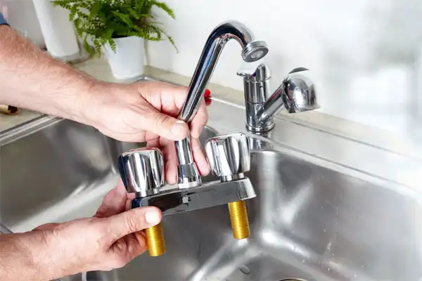 Hands are installing a dual-handle kitchen faucet above a sink, with a single-handle faucet visible in the background.