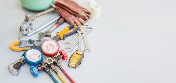 A collection of air conditioning repair tools, including wrenches, pliers, gloves, and a manifold gauge set, arranged on a light surface in Monticello, MN.